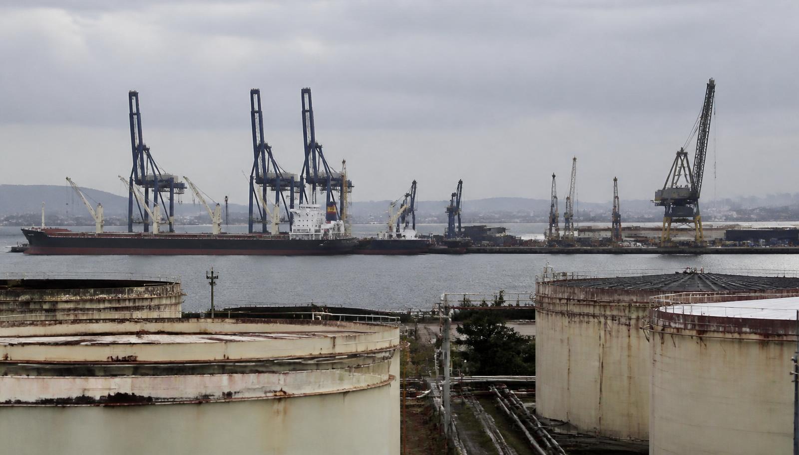 Terminal portuário industrial na Bahia com guindastes, navio atracado e tanques de armazenamento em primeiro plano sob céu nublado
