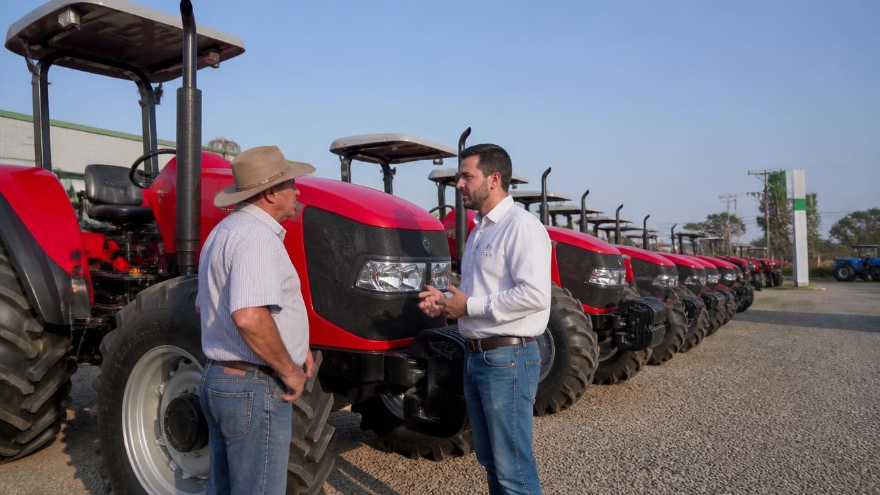 Produtor rural conversando com vendedor em pátio de concessionária de máquinas agrícolas com tratores enfileirados sob céu limpo no interior da Bahia